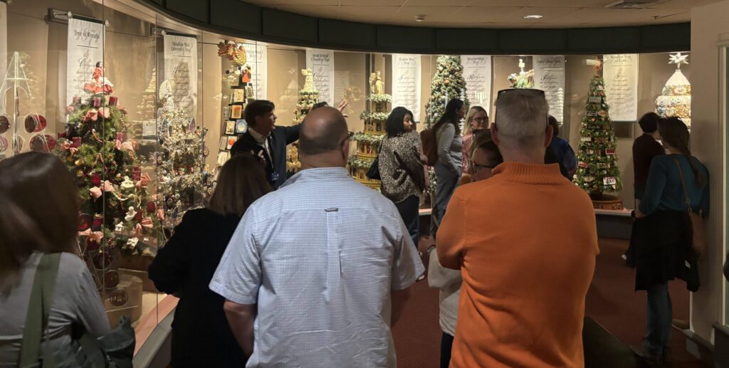 Members of Artisan Learning staff are listening to a tour guide at the Hallmark headquarters in Kansas City, Missouri, while looking a themed Christmas trees.
