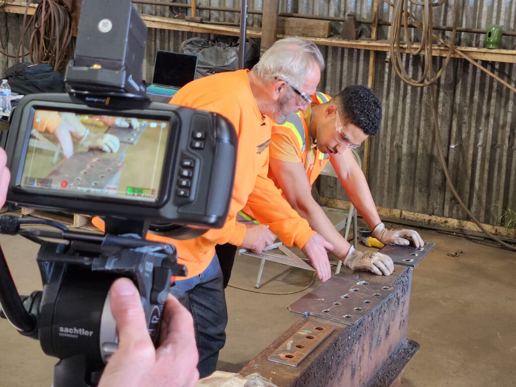 A videographer films a trainer and a trainee in steel fabrication shop.