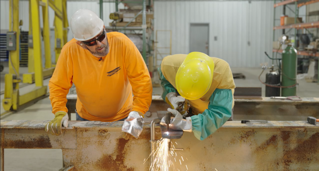 A trainer observes as a new steel worker welds.