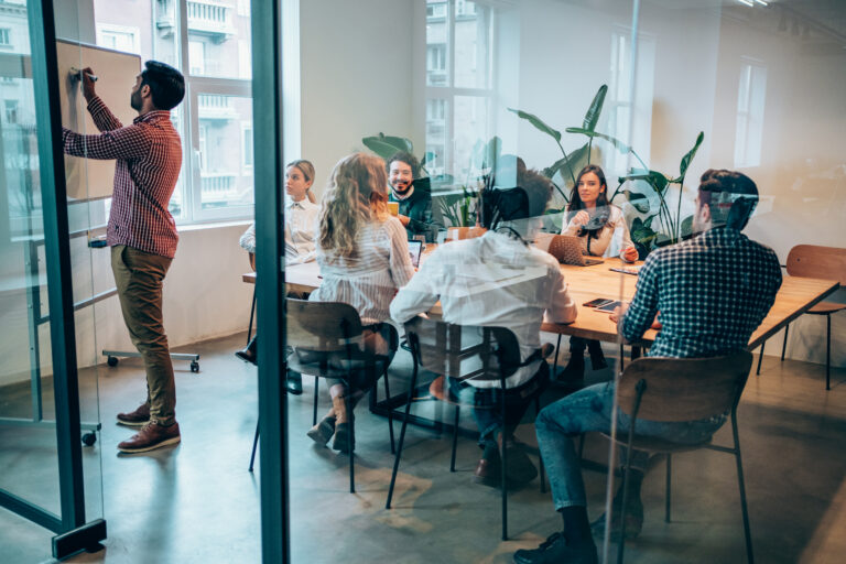 Businessman is writing on the whiteboard. Business people watching a presentation on the whiteboard. Multi-ethnic group of creative people on a business meeting in modern office. Creative team sitting on a conference table in board room. The view is through glass.
