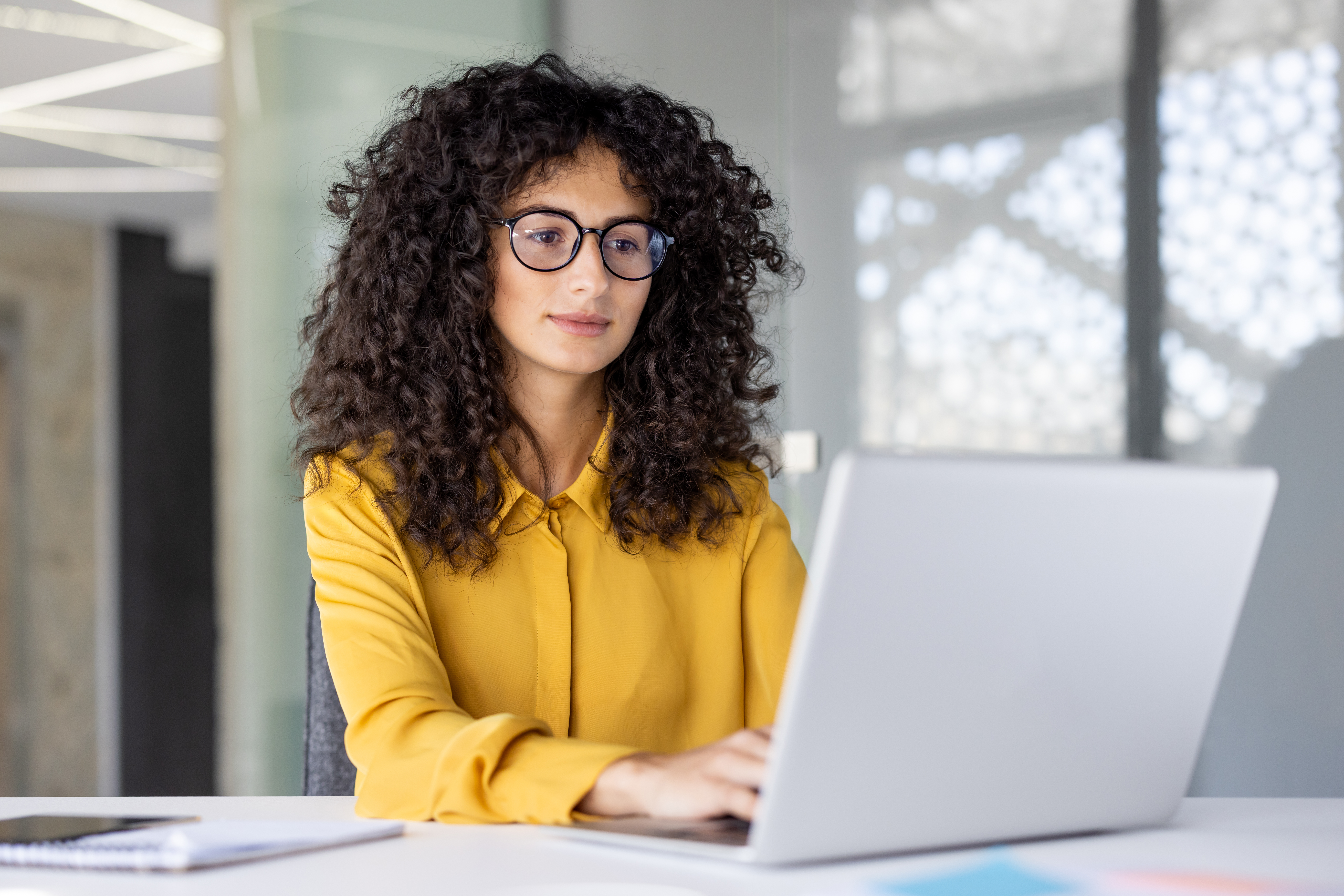 A woman with curly hair, wearing glasses and a yellow shirt, works on a laptop in an office setting. She appears focused and engaged in her tasks at the white desk.